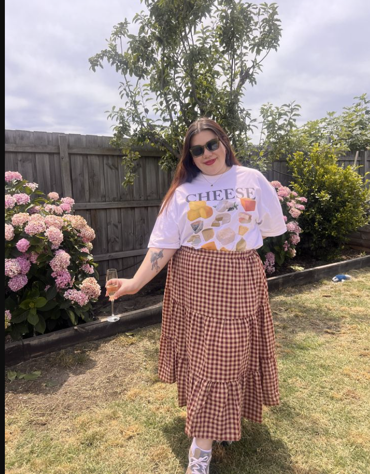 A plus size woman wearing White graphic cheese print t-shirt Brown gingham tiered midi skirt Black sunglasses White sneakers