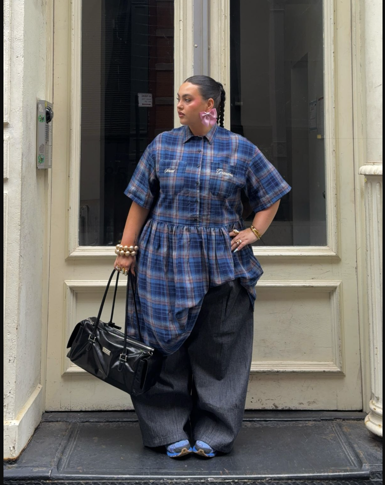 A plus size woman wearing Blue plaid short-sleeve shirt Dark wide-leg pants Pink hair bow Black handbag Gold bracelets and rings Blue patterned shoes