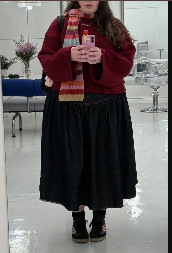 A plus size woman wearing Red oversized knit sweater Multi-colored striped scarf Dark denim midi skirt Black sneakers with pink accents Gold necklaces