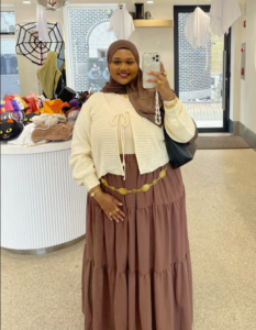 A plus size woman wearing Brown hijab White open-knit cardigan Cream top Brown tiered maxi skirt Gold statement belt Black handbag Gold jewelry