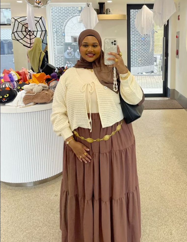 A plus size woman wearing Brown hijab White open-knit cardigan Cream top Brown tiered maxi skirt Gold statement belt Black handbag Gold jewelry