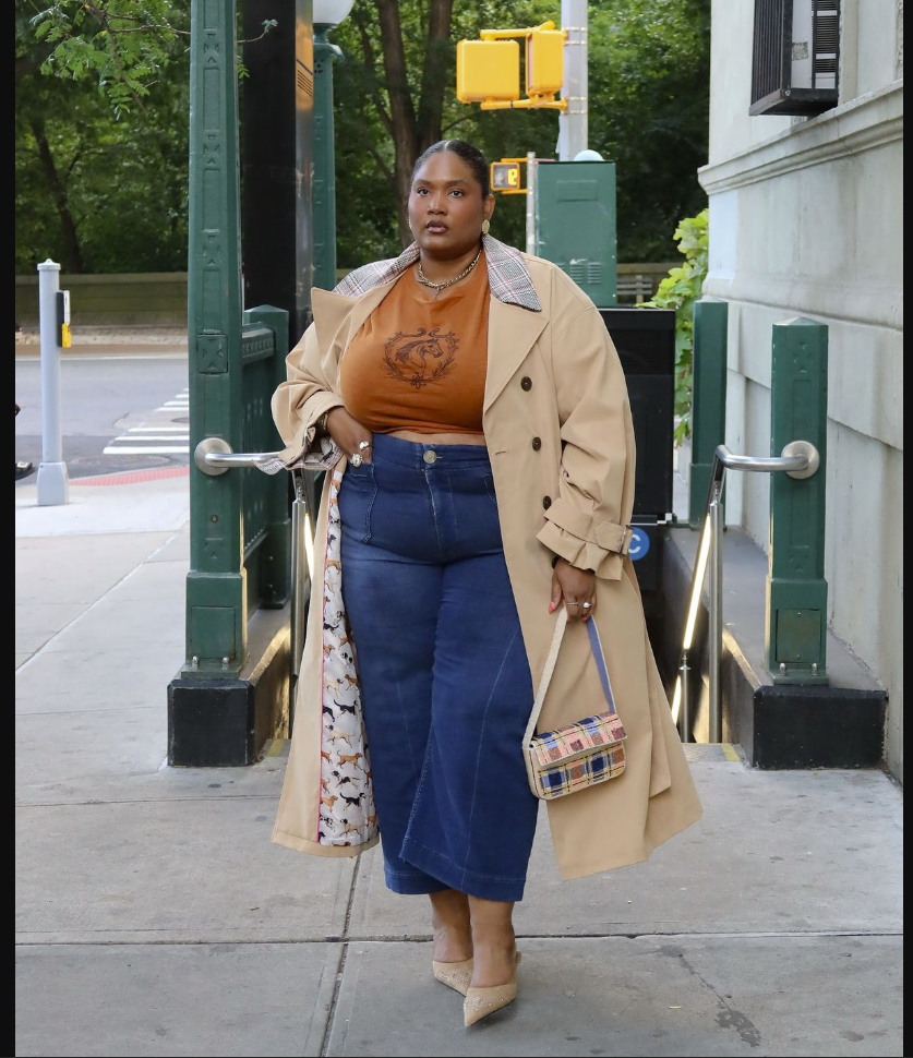 A plus size woman wearing Beige trench coat Orange graphic t-shirt Blue denim midi skirt Plaid crossbody bag Layered necklaces Pointed heels
