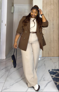 A plus size woman Brown double-breasted blazer Cream ribbed tank top Cream wide-leg trousers Brown belt with silver buckle Black and white two-tone sneakers Black handbag Long wavy braids in high ponytail Gold jewelry and watch