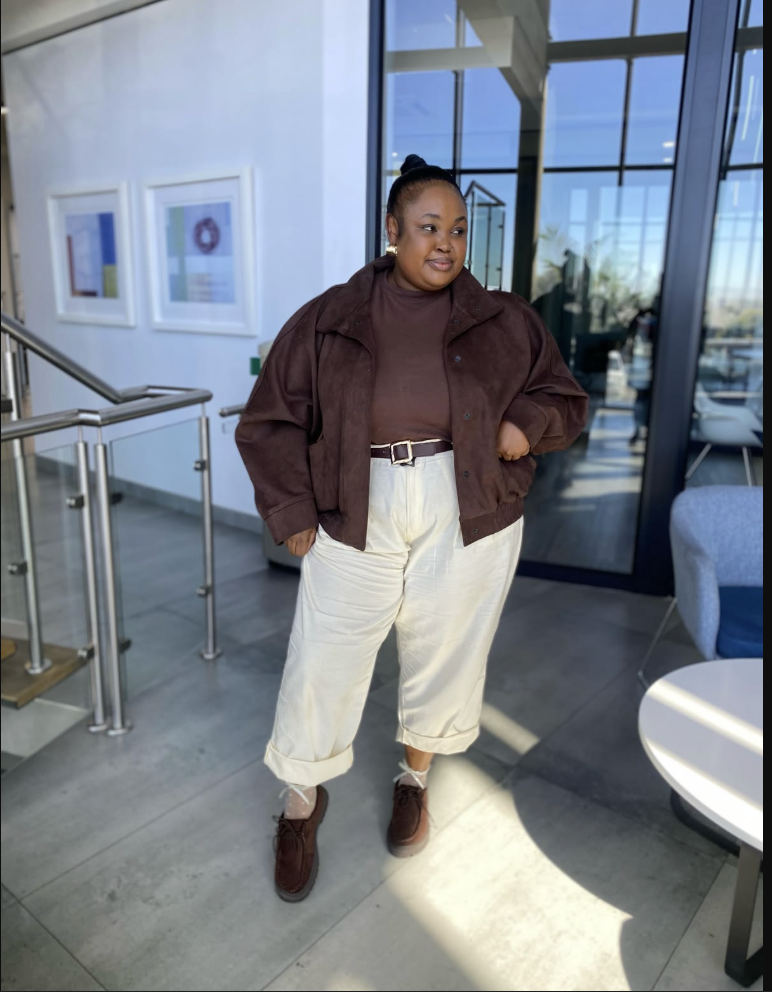 A plus size woman wearing Brown suede-style cropped jacket Brown turtleneck top Off-white cuffed wide-leg trousers Brown leather belt with silver buckle Brown suede lace-up shoes Gold earrings Hair in sleek bun