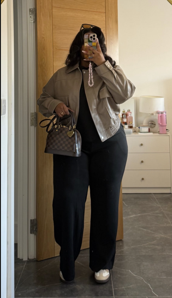 A plus size woman wearing Beige faux leather cropped jacket Black basic t-shirt Black wide-leg trousers White sneakers Brown checkered handbag with gold hardware Pearl phone strap Sunglasses on head Gold jewelry