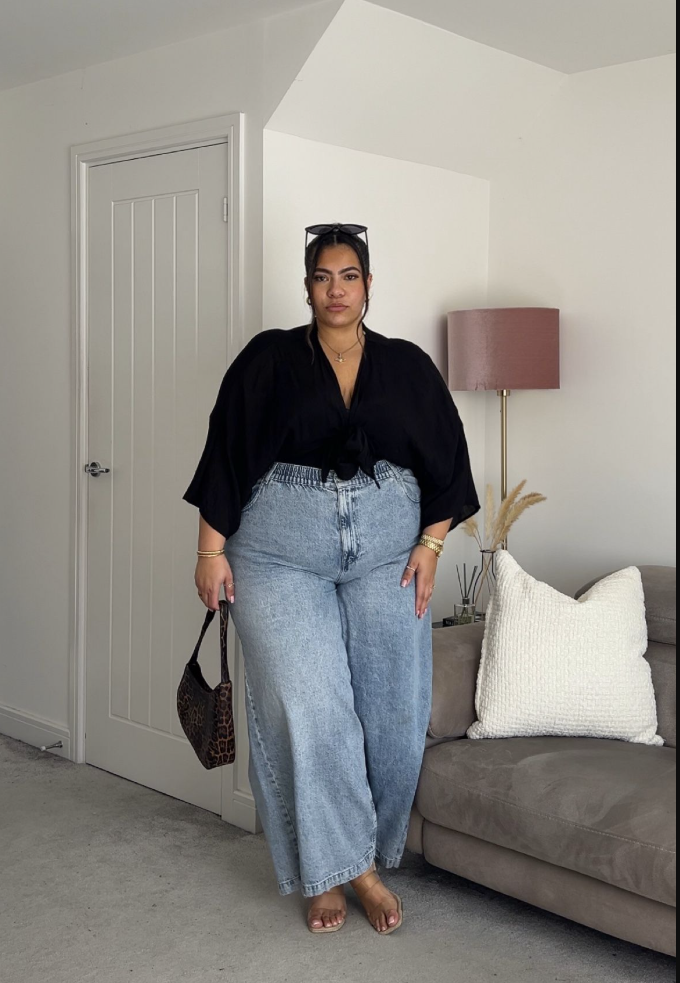 A plus size woman wearing Black tie-front kimono-style blouse
Light-wash wide-leg jeans
Leopard-print handbag
Sunglasses on head
Gold jewelry
Bare feet with sandals