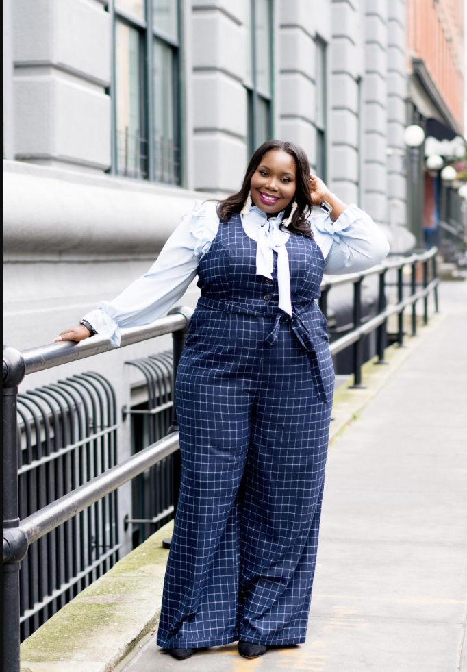 A plus size woman wearing Navy blue plaid wide-leg jumpsuit
Light blue ruffled button-up shirt underneath
White bow tie at neck
Black shoes
Gold jewelry