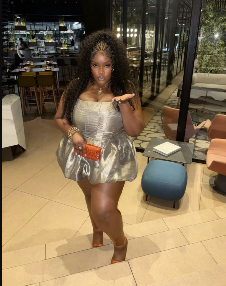A plus size woman wearing Silver satin strapless corset mini dress with bubble hem Orange glitter clutch Gold multi-layered necklace with pendant Gold statement earrings Gold multi-row bracelet Clear/orange strap high heels