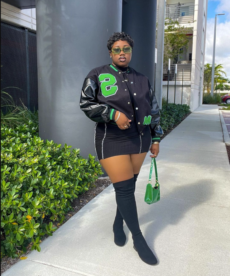 A plus size woman weaRING Black and green varsity bomber jacket with large "S" letter Black ribbed mini skirt with white side stripes Black over-the-knee boots Green mini shoulder bag Green-tinted rectangular sunglasses Silver hoop earrings Silver bracelet