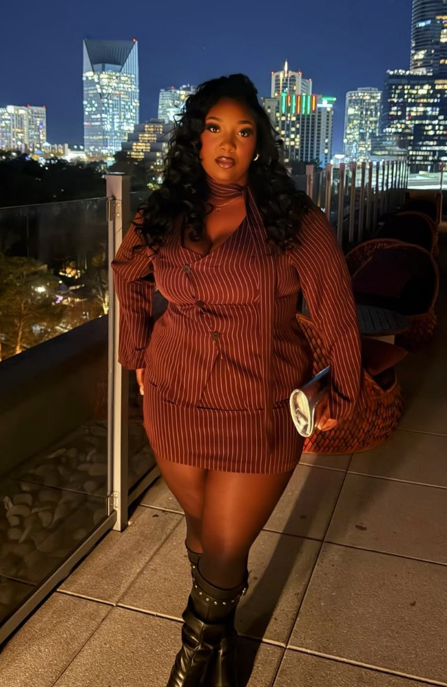 A plus size woman wearing Rust brown pinstripe blazer mini dress Black knee-high studded boots Gold hoop earrings Gold layered necklace Woven clutch or phone