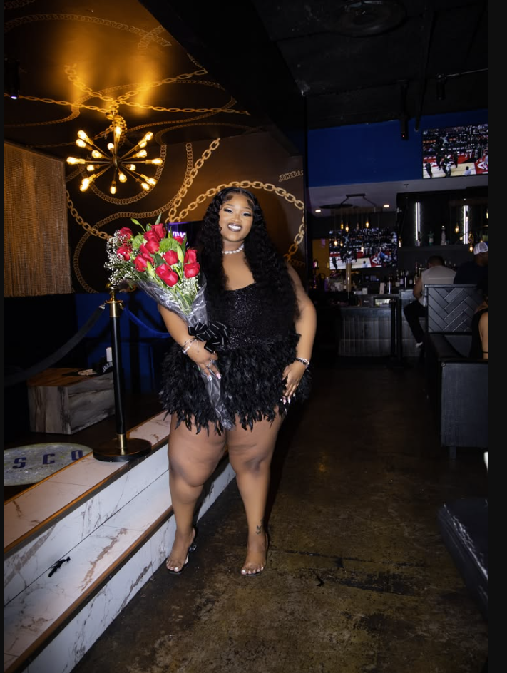 A plus size woman wearing Black sequin top with feather trim mini dress
Clear high heels
Pearl necklace
Silver bracelets
Bouquet of red roses