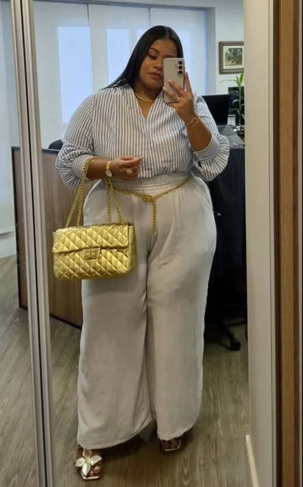 A plus size woman wearing Striped button-down shirt White wide-leg trousers Gold chain-link belt Gold quilted shoulder bag Gold necklace Gold watch Gold hoop earrings Metallic flower sandals