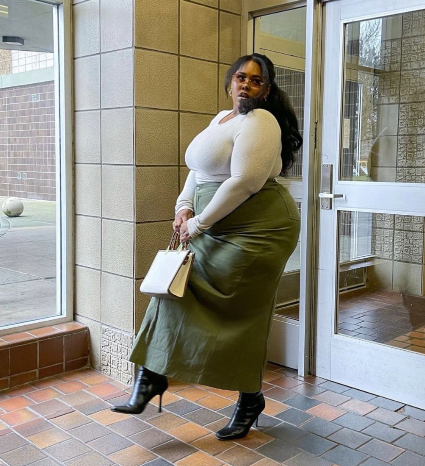 A plus size woman wearing White long-sleeve bodysuit Olive green midi skirt Black pointed-toe heeled boots White structured handbag Gold hoop earrings Thin-rimmed glasses