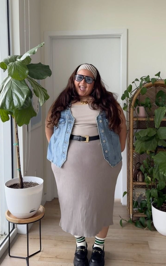 A plus size woman wearing Beige ribbed tank top Light-wash denim vest Beige midi skirt Black leather belt with gold buckle Striped knit headband Black chunky loafers Striped socks