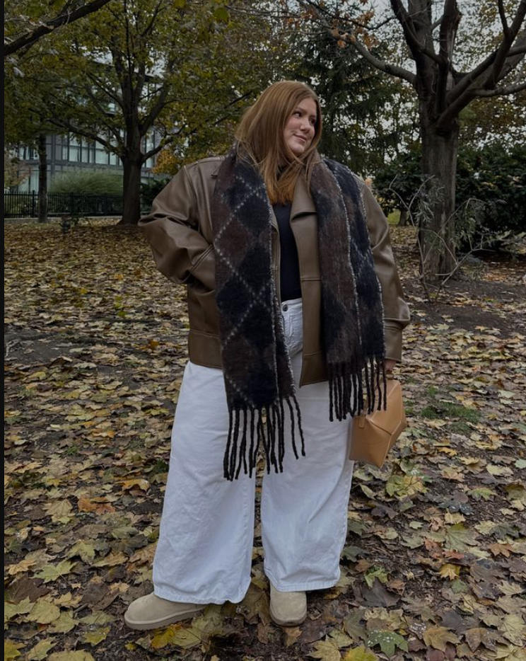 A plus size woman wearing Brown leather bomber jacket Plaid fringe scarf Black top White wide-leg jeans Beige suede boots Tan structured handbag