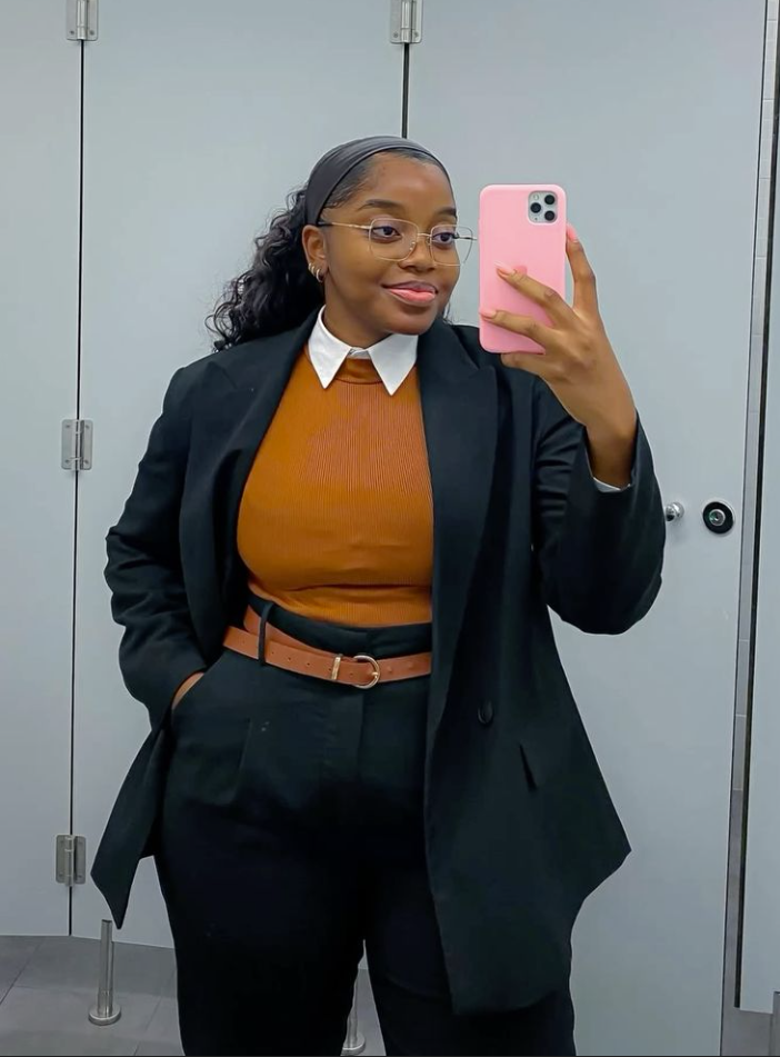 A plus size woman wearing Black oversized blazer Orange ribbed sweater White collared shirt Black tailored trousers Brown leather belt Thin-rimmed glasses