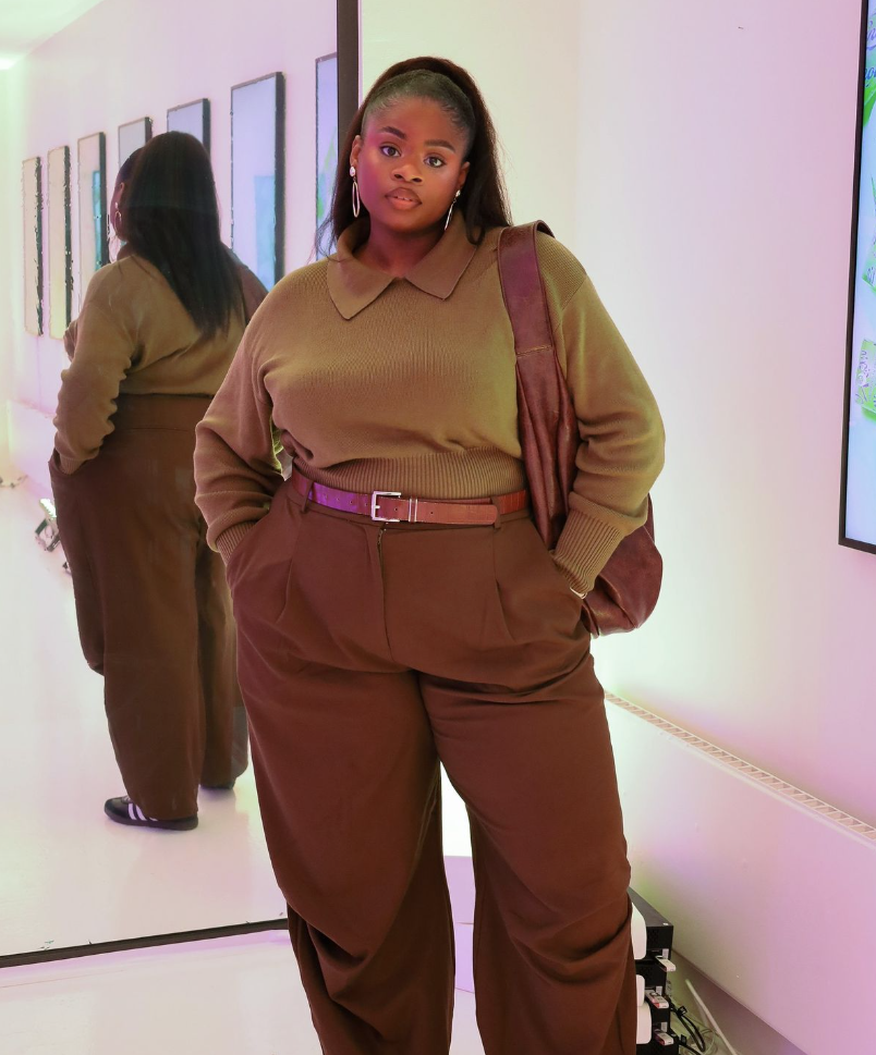 A plus size woman wearing Brown collared sweater Brown wide-leg trousers Brown leather belt Brown shoulder bag Gold hoop earrings