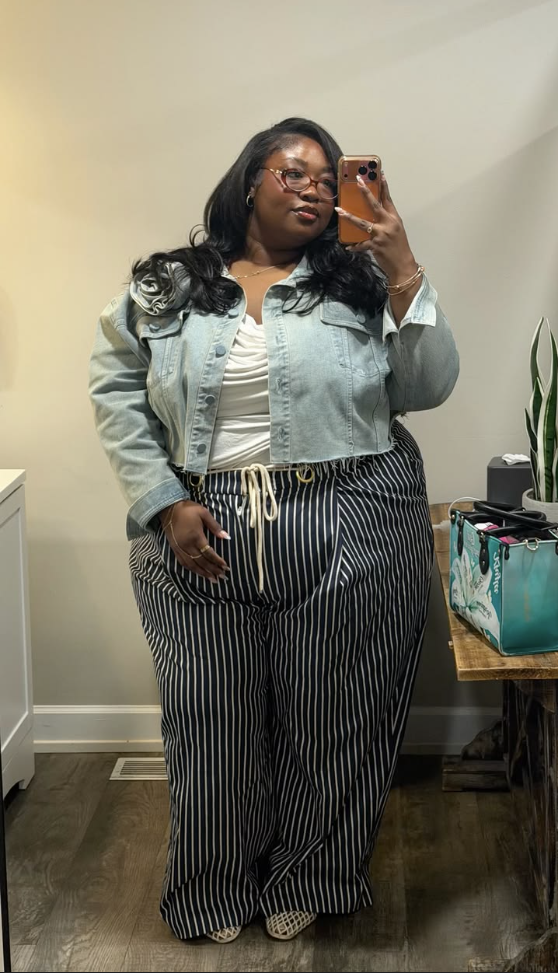 A plus size woman wearing Light blue denim jacket
White ribbed tank top
Black and white striped wide leg pants
Gold chain belt
Patterned flat shoes