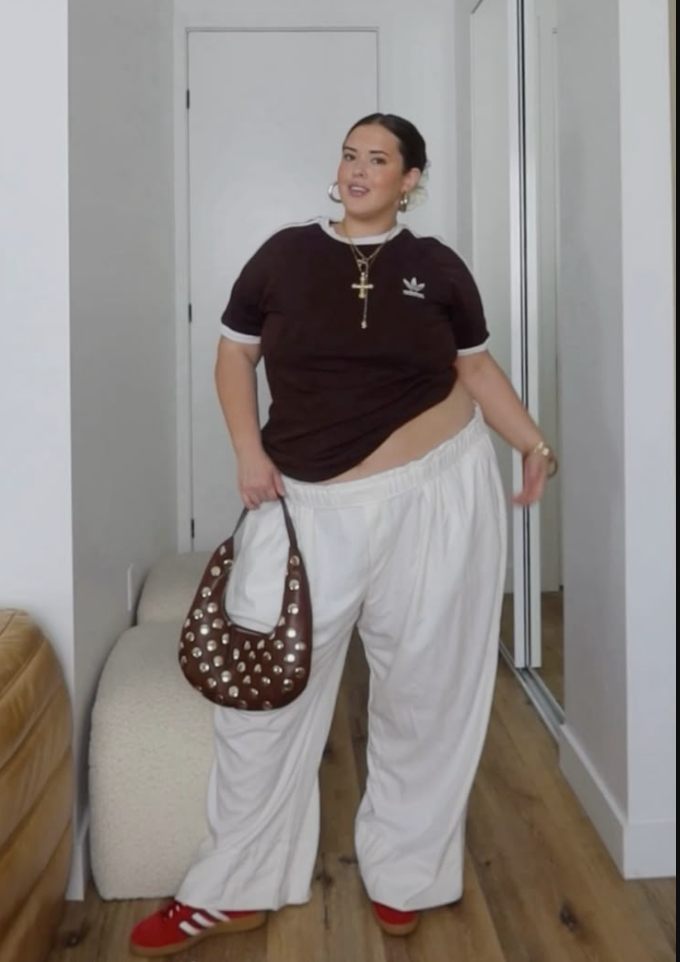 A plus size woman wearing Brown Adidas t-shirt
White track pants
Red sneakers
Brown studded shoulder bag
Gold cross necklace
