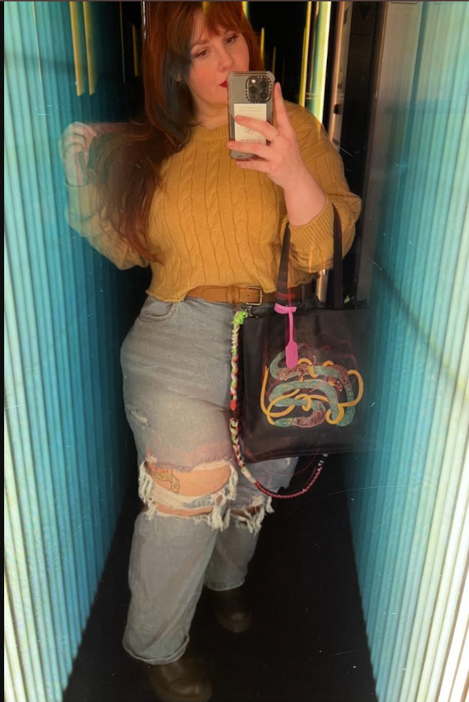 A plus size woman wearing Mustard yellow cable knit sweater
Light-wash ripped jeans
Brown leather belt
Graphic tote bag