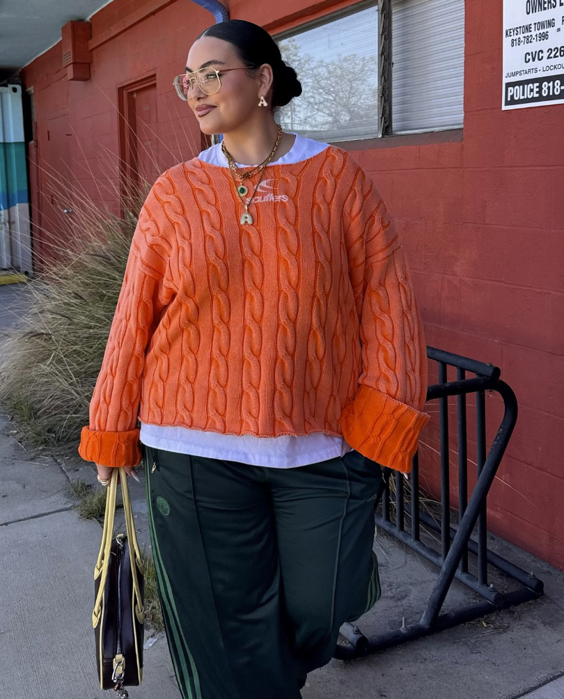A plus size woman wearing Orange cable knit sweater
White layered top
Dark green track pants with stripes
Gold layered necklaces
Handbag