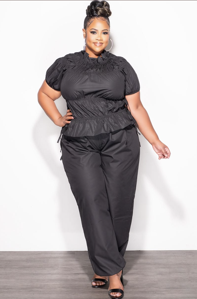 A plus size woman wearing Black ruched puff-sleeve blouse Black high-waisted wide-leg trousers Black peplum waist detail top Large silver hoop earrings Black open-toe heeled sandals