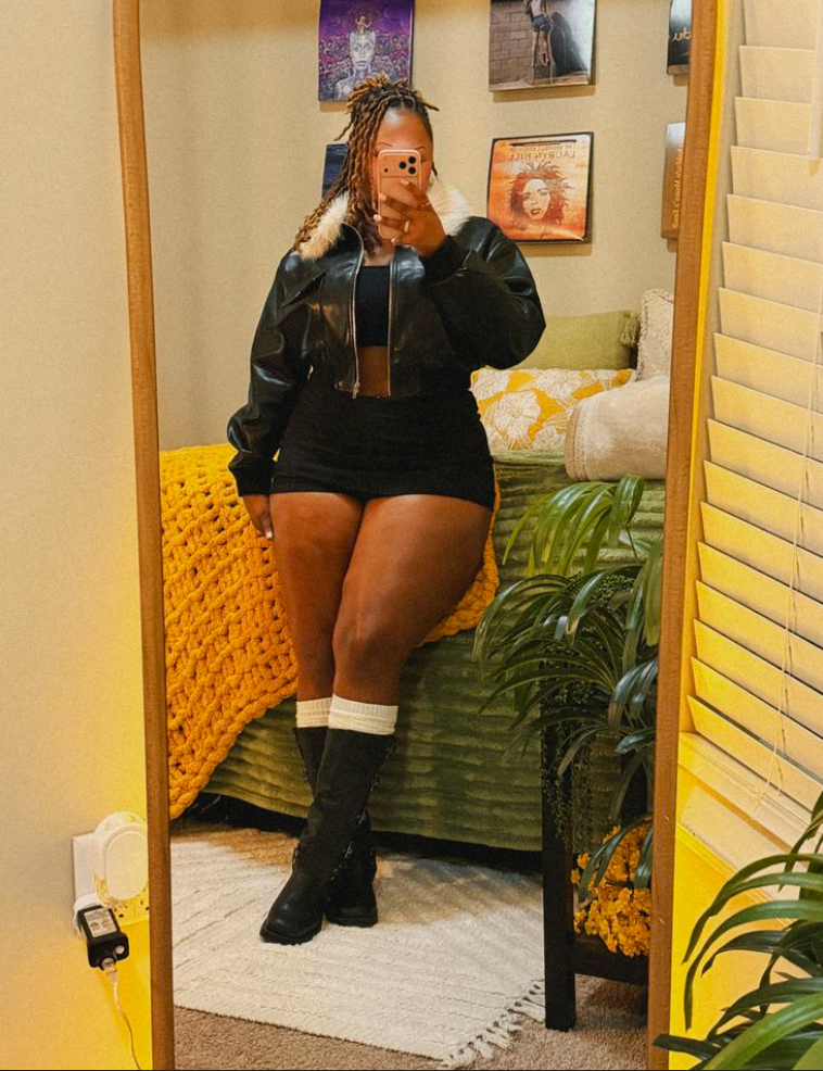 A plus size woman wearing Black cropped faux leather jacket with white fur collar Black crop top Black mini skirt Black knee-high boots White slouchy crew socks