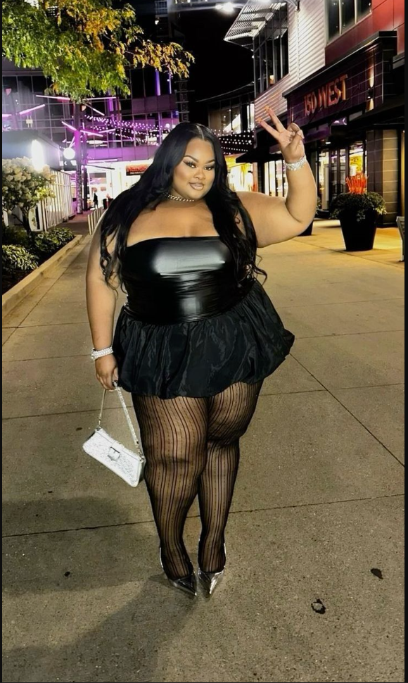 A plus size woman wearing Black strapless leather-like bandeau top
Black ruffled mini tutu skirt
Black fishnet tights
Silver metallic pointed-toe heels
White chain-strap crossbody bag
Silver chain necklace
Silver tennis bracelet