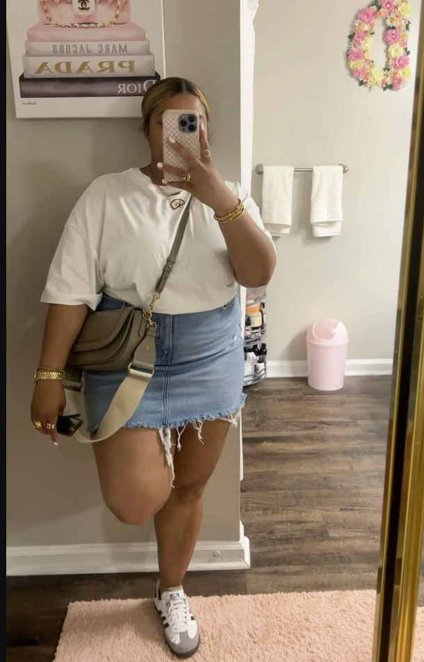 A plus size woman wearing White oversized t-shirt Light-wash denim mini skirt with frayed hem Taupe crossbody bag White and gray sneakers Gold bangles Gold rings