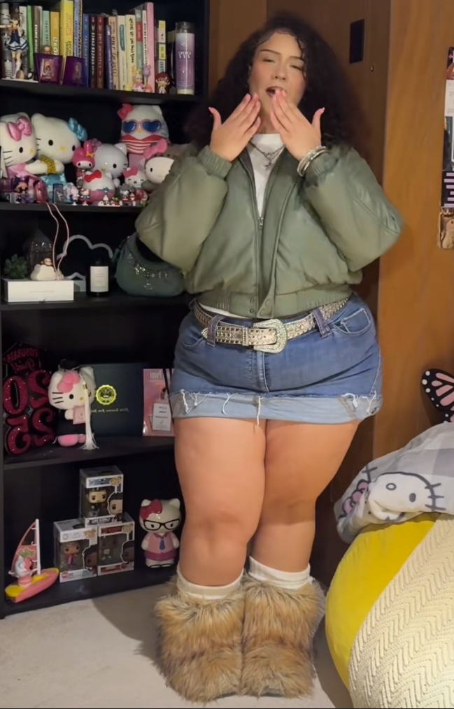 A plus size woman wearing Olive green bomber jacket
White t-shirt
Light-wash denim mini skirt
Sparkly statement belt
Beige furry boots
Silver layered necklaces
Silver bracelets