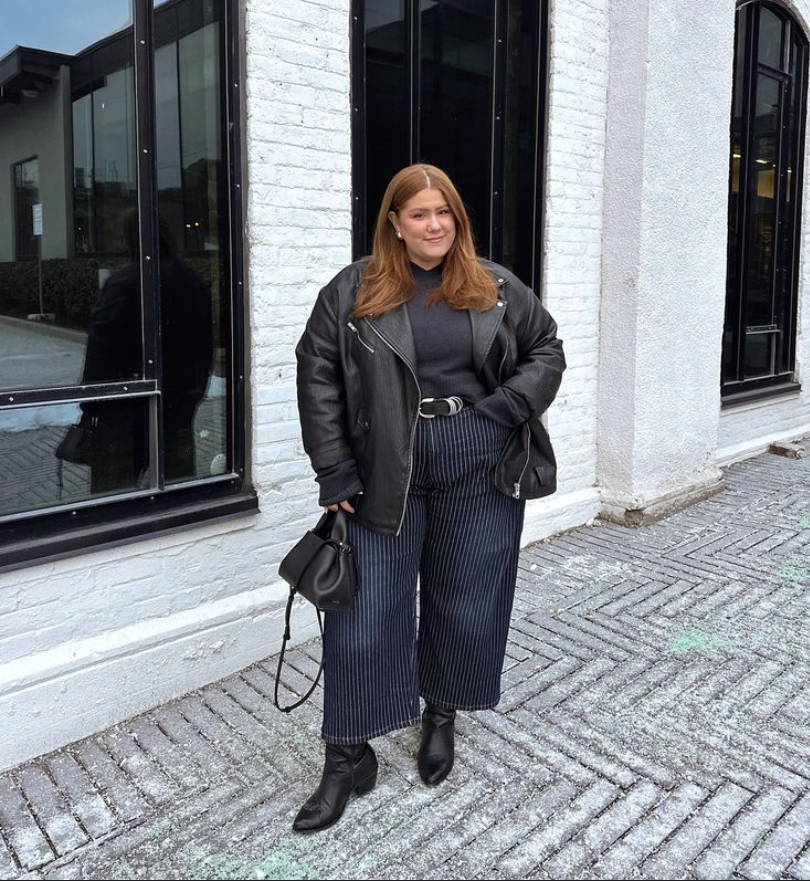 A plus size woman wearing Black leather moto jacket
Dark gray turtleneck sweater
Navy pinstripe wide-leg trousers
Black ankle boots
Black crossbody bucket bag
Silver belt