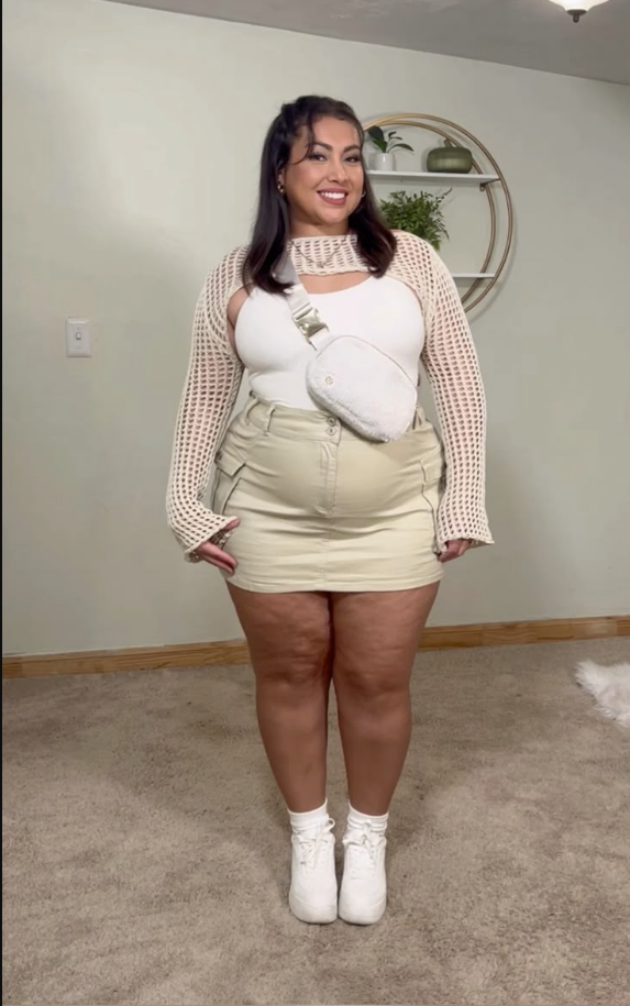 A plus size woman wearing White ribbed tank top Cream open-knit long-sleeve cover-up Cream high-waisted cargo mini skirt White lace-up sneakers White fanny pack Pearl choker necklace
