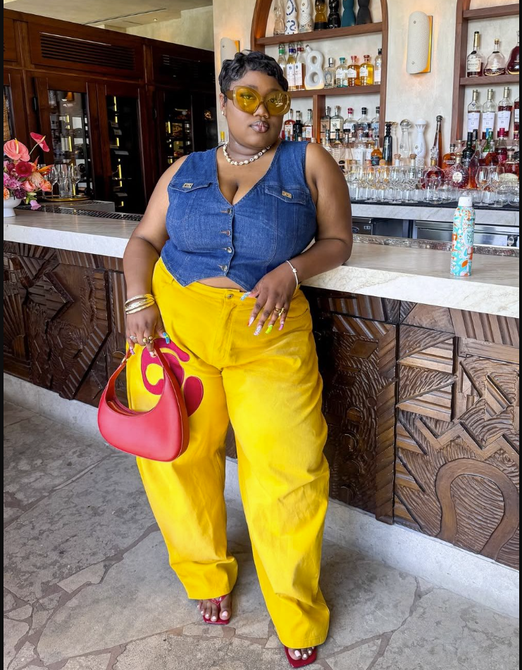 A plus size woman wearing Blue denim sleeveless button-up vest
Bright yellow wide-leg trousers
Red curved handbag
Yellow-tinted sunglasses
Pearl necklace
Gold bracelets and rings
Red heeled sandals