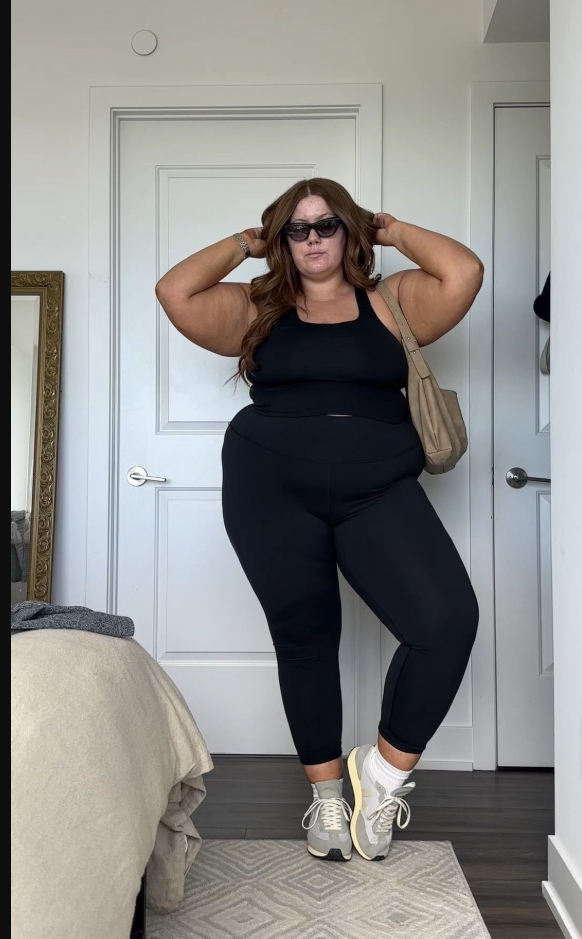 A plus size woman wearing Black halter crop top
Black high-waisted leggings
Grey and white chunky sneakers
Beige shoulder bag
Black sunglasses