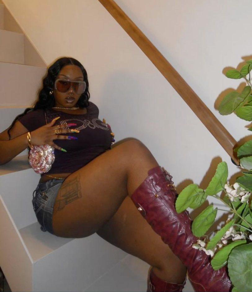 A plus size woman wearing Purple rhinestone graphic baby tee Light-wash denim mini skirt Burgundy snakeskin knee-high boots Gold layered necklaces Gold bangles Oversized shield sunglasses Sequin disco ball handbag