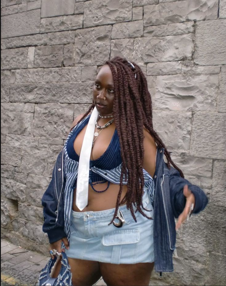 A plus size woman wearing Blue striped halter bralette Light-wash denim mini skirt with pockets Blue denim jacket White necktie Layered silver necklaces Silver rings Denim handbag 