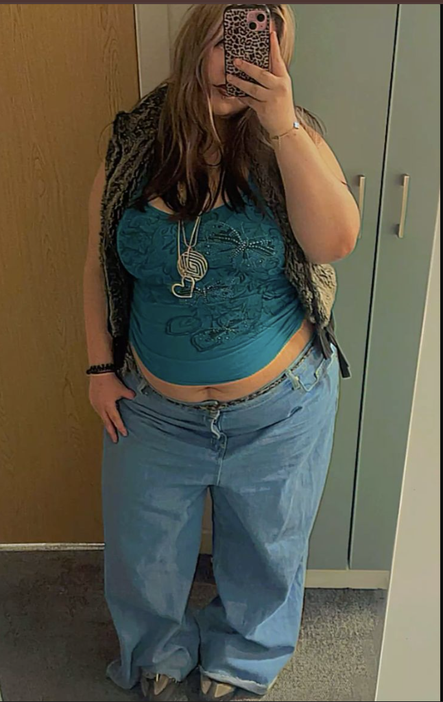 A plus size woman wearing Teal embroidered baby tee Light-wash baggy jeans Black faux fur vest Layered necklaces with heart pendant Leopard print phone case