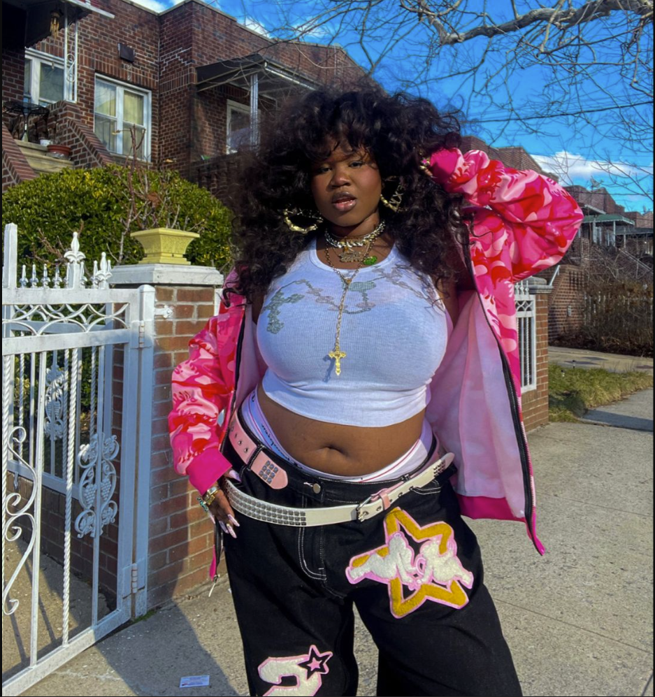 A plus size woman wearing Light blue graphic baby tee Black cargo-style pants with star patches Pink and white camo zip-up jacket White studded belt Gold layered necklaces with cross pendant Large gold hoop earrings
