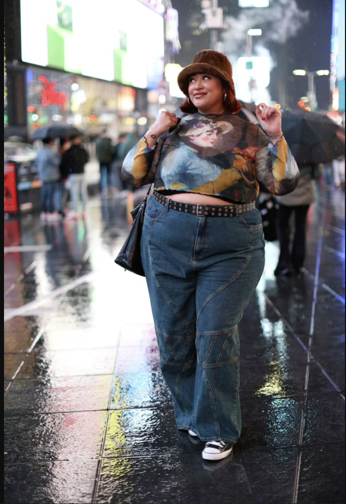 A plus size woman wearing Multi-color graphic mesh long-sleeve crop top Dark blue wide-leg cargo jeans Brown fuzzy bucket hat Studded black belt Black shoulder bag