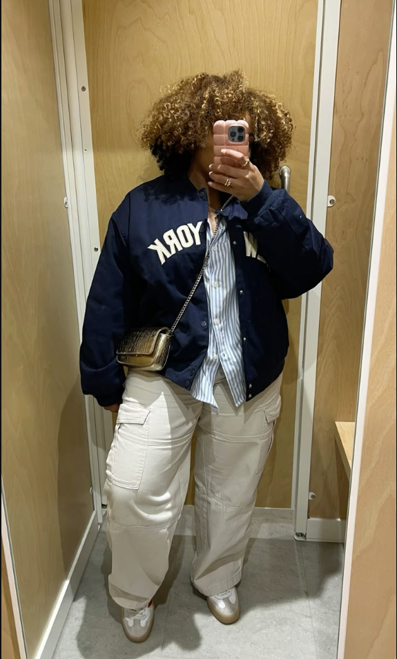 A plus size woman wearing Navy blue varsity bomber jacket Striped button-down shirt White high-waisted cargo pants Gold chain crossbody bag White sneakers Gold ring Silver necklace
