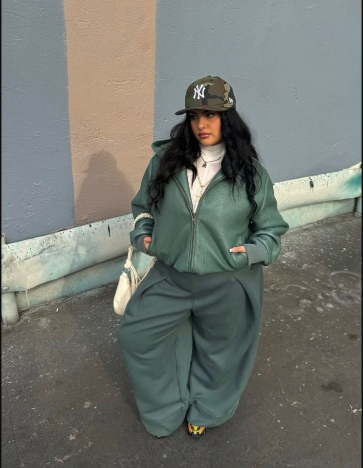 A plus size woman wearing Green faux leather zip-up hoodie jacket Camouflage baseball cap White turtleneck Matching green wide-leg pants White shoulder bag Patterned sneakers