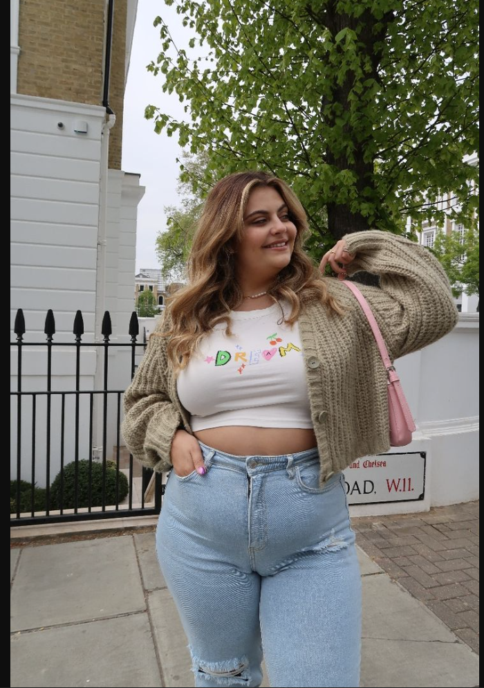 A plus size woman wearing Beige knit cardigan
White graphic crop top
Light blue distressed jeans
Pink shoulder bag
