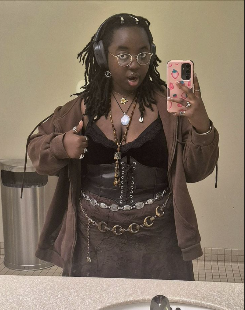 A plus size woman wearing Brown zip-up hoodie jacket Black lace bralette Black corset top Brown patterned midi skirt Silver and gold layered chain belts Multiple layered necklaces Over-ear headphones (as accessory) Silver hoop earrings