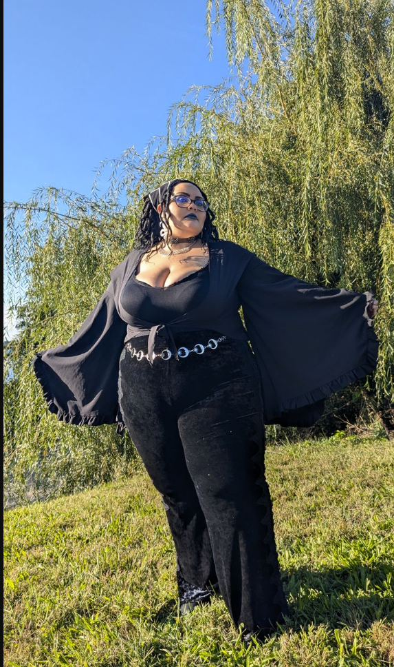 A plus size woman wearing Black wide bell sleeve wrap top Black velvet wide-leg pants Silver chain link belt Black choker