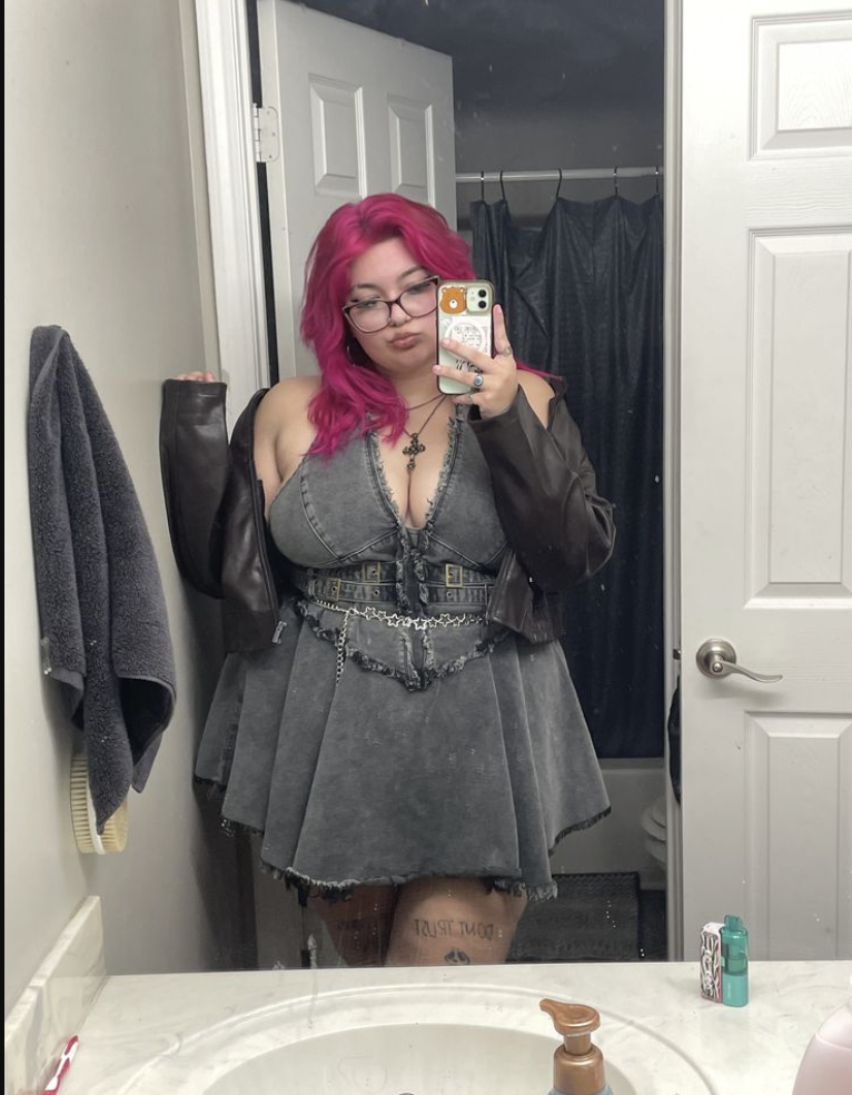A plus size woman wearing Gray acid-wash denim halter dress Black leather jacket Silver chain belt with hardware Layered necklaces