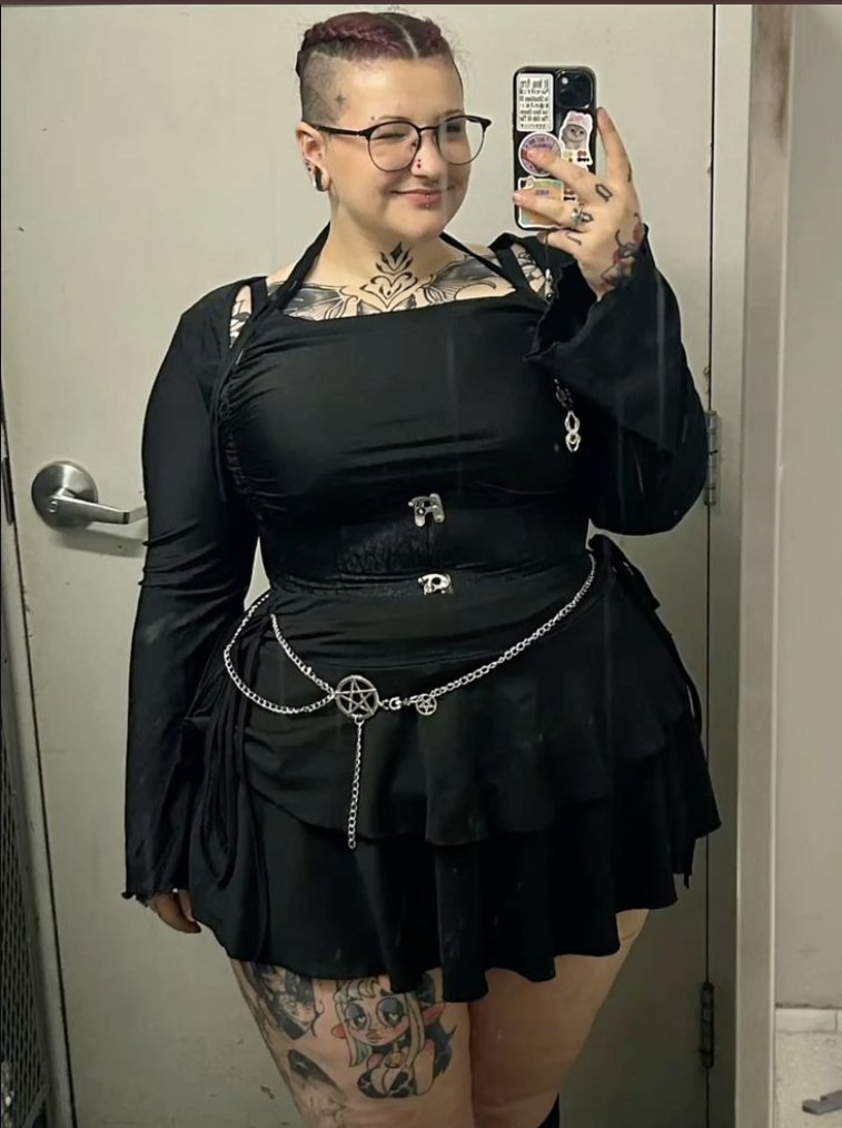 A plus size woman wearing Black long sleeve ruched top with harness straps Black tiered ruffle mini skirt Silver chain belt with pentagram Silver earrings