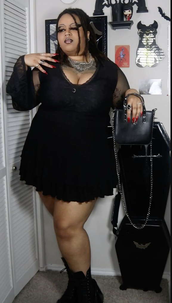 A plus size woman wearing Black lace sleeve mini dress Silver chain choker Black platform combat boots Black spiked handbag