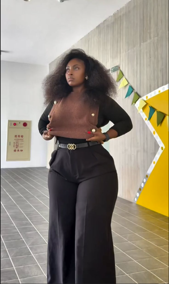 A plus size woman wearing Black long sleeve top
Brown button-front knit vest
Black wide leg trousers
Black belt with gold buckle